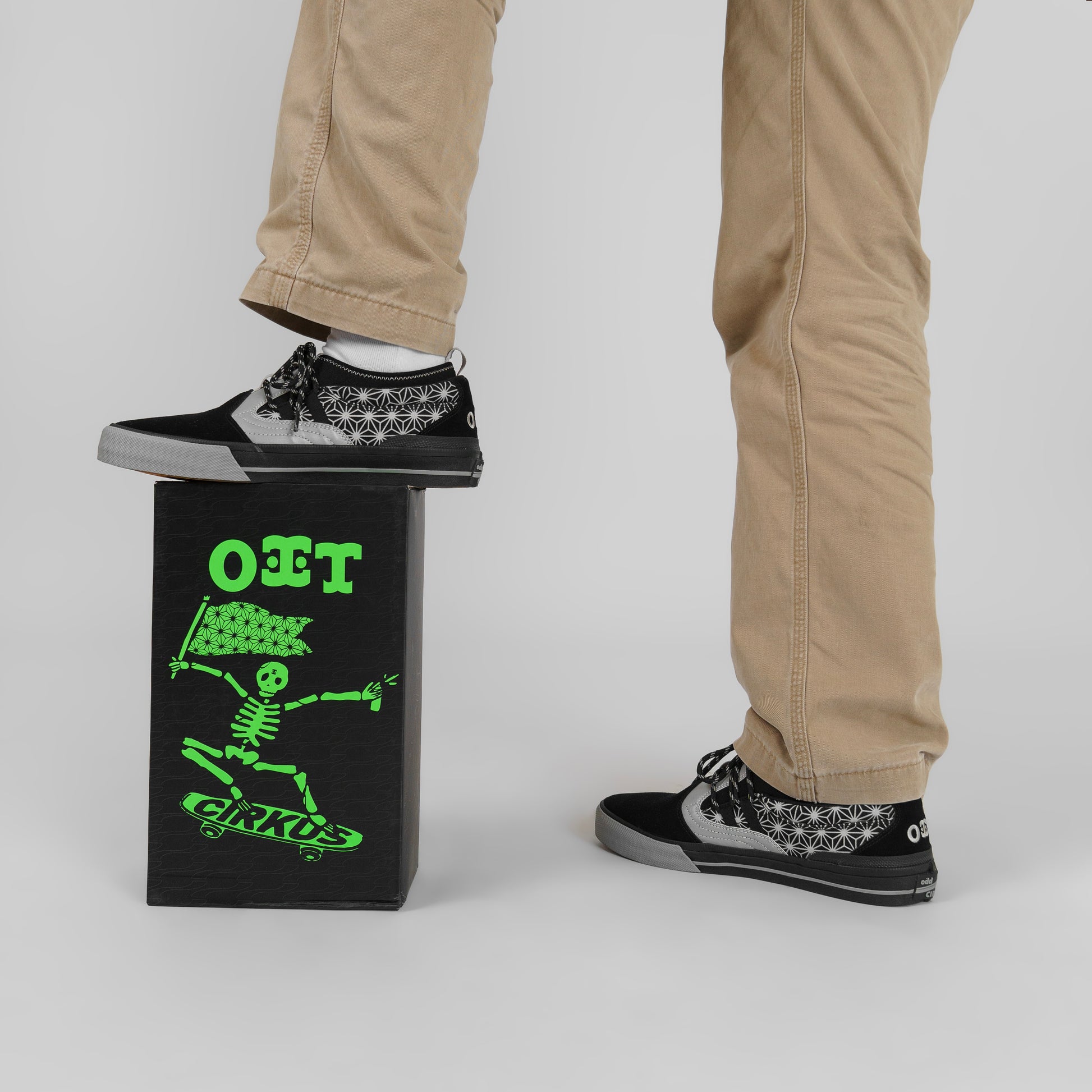Person wearing black and white patterned sneakers with beige pants, standing next to a black box with green text and graphics.